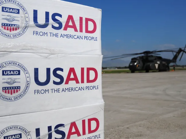 Boxes of aid from USAID are stacked on a tarmac near a US military helicopter